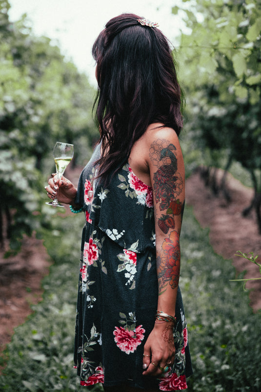 Frau in dunklem Blumenkleid und einem Glas Wein in der Hand schaut auf eine Weinplantage.